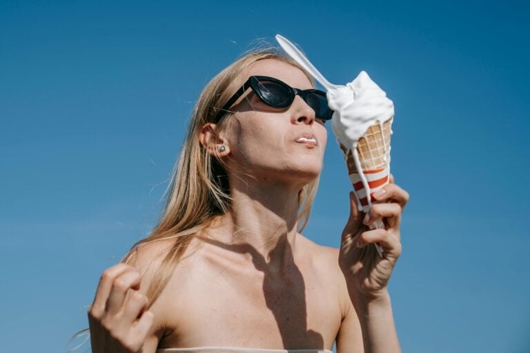 Une femme blonde est photographiée par un jour ensoleillé. Le ciel est bleu, elle mange une glace à la vanille qui fond et coule sur sa main et son menton.