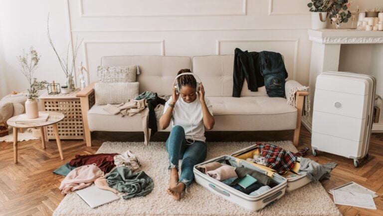 Une femme, écouteurs sur les oreilles, est assise dans son salon, entourée d'affaires. Elle prépare ses valises pour les vacances, et sélectionne les meilleurs sextoys à emporter.