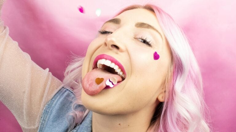 Une femme aux longs cheveux colorés est heureuse, la bouche grande ouverte, la langue sortie. L'atmosphère pop et léger de l'image illustre parfaitement notre propos sur la gorge profonde.