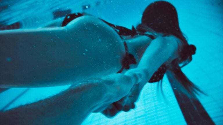 Une femme nage sous l'eau dans la piscine, prenant par la main un partenaire pour vivre une historie érotique dans les douches.