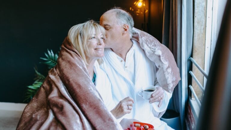 Un couple s'enlace et s'embrasse devant une fenêtre, enroulé dans une couette. La femme a transitionné vers la ménopause.