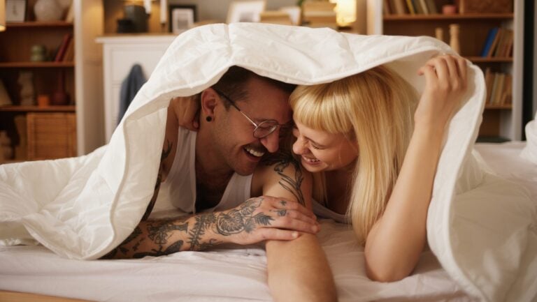 Un couple allongé sur le ventre sur leur lit est prêt à vivre une histoire érotique. Ils rient en se regardant, la couette posée au dessus de leur tête.